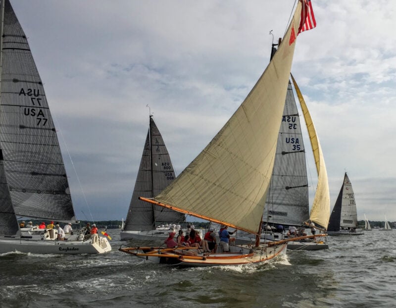 Several sailboats, including a wooden boat with tan sails and people onboard, race closely together on a slightly choppy body of water under a cloudy sky, with land—and nearby Annapolis condos for sale—visible in the distance.