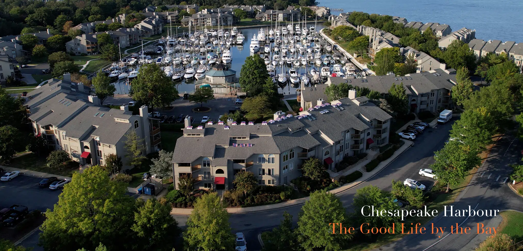 Aerial view of Chesapeake Harbour marina with boats docked, surrounded by tree-lined residential buildings near the bay. The text reads, Chesapeake Harbour - Annapolis Condos for Sale: The Good Life by the Bay.