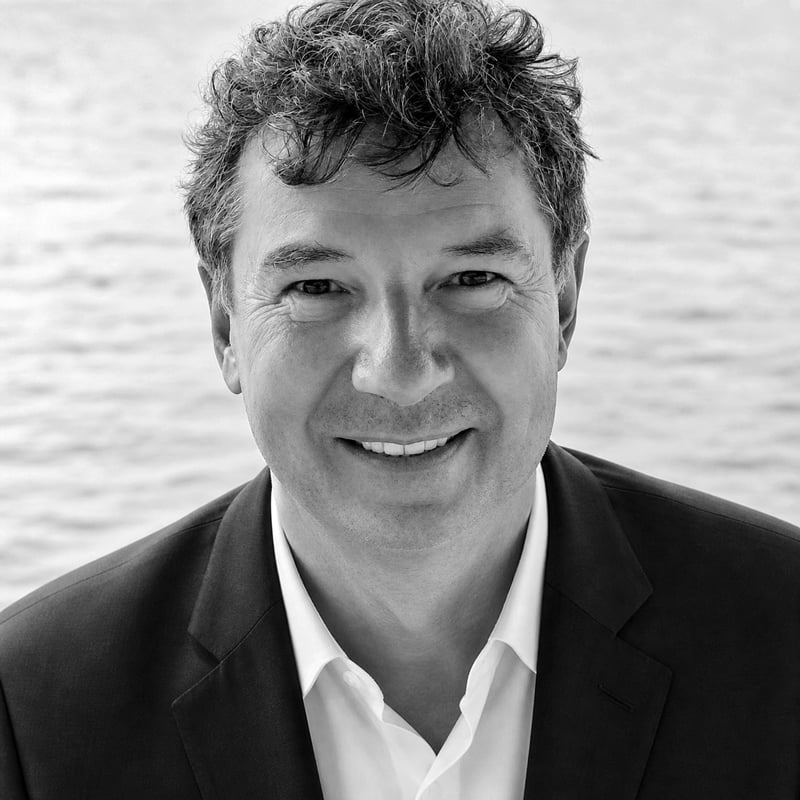 A man with wavy dark hair, wearing a suit jacket and open-collared shirt, smiles at the camera. Behind him, water glistens—perfect for showcasing Annapolis Condos for Sale. The image is in black and white.