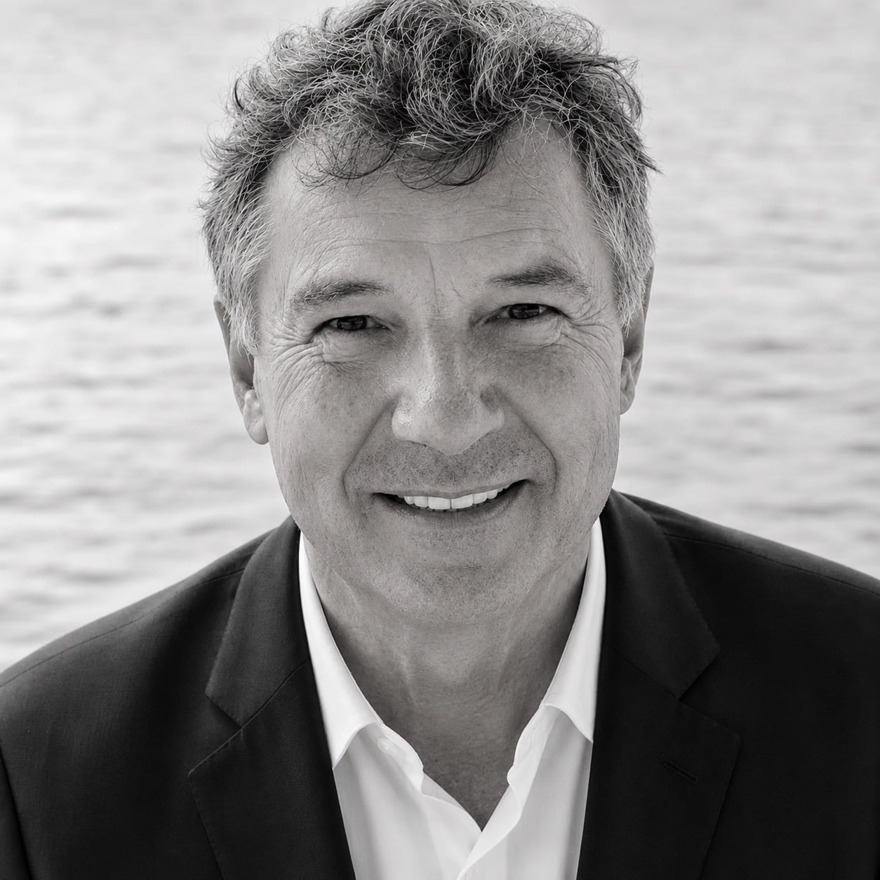 Smiling man with gray hair wearing a dark blazer and white shirt, standing outdoors with a blurred body of water in the background, embodying the relaxed elegance found near Annapolis Condos for Sale.