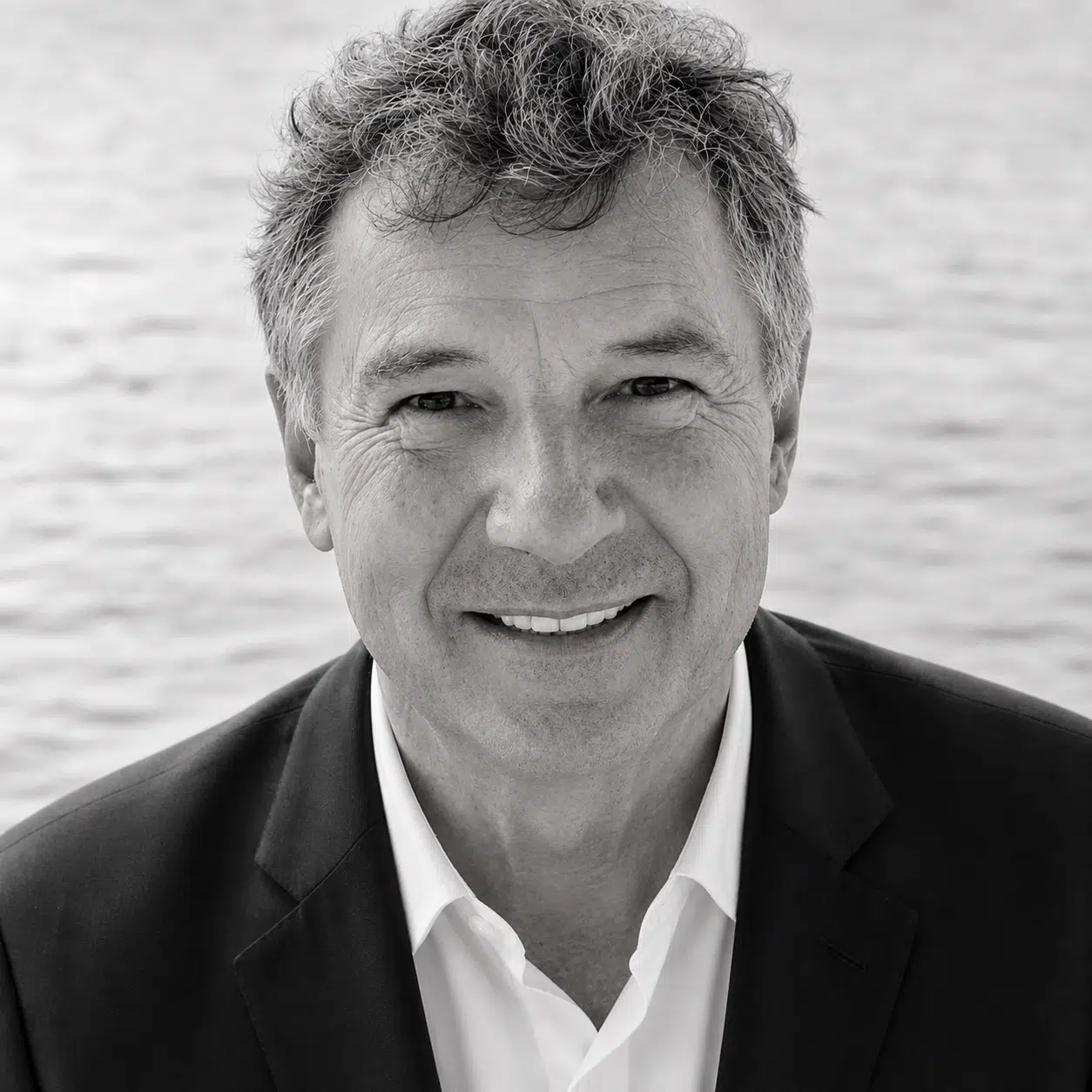 Smiling man with gray hair wearing a dark blazer and white shirt, standing outdoors with a blurred body of water in the background, embodying the relaxed elegance found near Annapolis Condos for Sale.